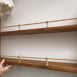 Puede incluir: Estantería de pared de madera de dos niveles con rieles y detalles de latón. Los estantes son de color marrón cálido, y los rieles de latón están soportados por postes decorativos. La estantería está montada en una pared blanca. El diseño es vintage y decorativo.