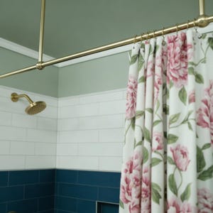 Könnte beinhalten: Ein Badezimmer mit einer goldenen Duschstange und Duschkopf. Ein floraler Duschvorhang mit rosa Pfingstrosen und grünen Blättern hängt an der Stange. Die Wände sind weiß gefliest mit einem blauen Fliesensockel und einer grünen Decke.