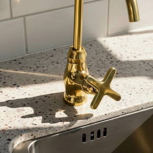 May include: A close-up of a gold-colored faucet with a cross-shaped handle. The faucet is installed on a speckled countertop next to a stainless steel sink. White rectangular tiles are in the background.