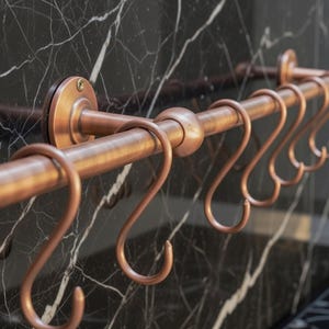 May include: A copper-coloured kitchen rail with six S-shaped hooks. The rail is mounted on a dark, veined surface. The design is simple and functional, suitable for hanging kitchen utensils or other items.