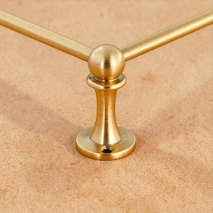 Könnte beinhalten: Nahaufnahme eines dekorativen Handtuchhalters in Messingoptik. Der Halter hat einen abgerundeten Sockel, einen zentralen Schaft und einen kugelförmigen Akzent, wo sich die beiden horizontalen Stangen treffen. Die Oberfläche hat einen warmen, metallischen Ton.