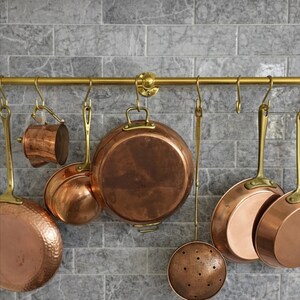 Puede incluir: Una colección de utensilios de cocina de cobre, que incluye ollas, sartenes y un colador, cuelga de una barra de latón contra una pared de azulejos grises. Los artículos de cobre tienen un tono marrón rojizo cálido.