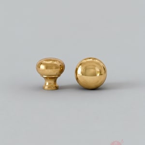 Könnte beinhalten: Zwei Messing-Schrankknöpfe. Einer hat eine ovale Form mit einem Sockel, der andere ist eine Kugel. Beide Knöpfe haben eine polierte, reflektierende Oberfläche und sind vor einem neutralgrauen Hintergrund platziert.