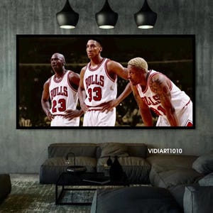 Puede incluir: Una impresi&oacute;n enmarcada con tres jugadores de baloncesto con camisetas blancas y rojas. Las camisetas muestran la palabra "Bulls" y los n&uacute;meros 23, 33 y 91. La impresi&oacute;n se muestra encima de un sof&aacute; gris oscuro.