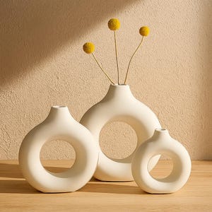 May include: Three off-white ceramic vases with a circular design. The vases are different sizes and arranged together. Yellow flowers are placed in the largest vase. The vases are on a wooden surface with a neutral background.