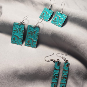 May include: Several pairs of turquoise and brown rectangular earrings with a floral design. The earrings have a distressed, textured appearance and silver-colored ear wires. The earrings are displayed on a gray fabric surface.