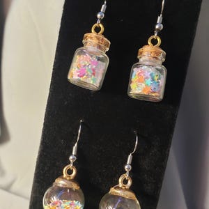 May include: Four pairs of earrings. Two pairs feature small glass bottles with cork stoppers, filled with colorful star-shaped confetti. The other two pairs are clear glass globes filled with similar confetti, all hanging from silver-toned hooks.