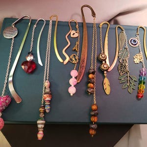 Op de afbeelding: Een collectie van negen metalen bladwijzers met diverse decoratieve bedels, waaronder een hart, een rode kristal, papieren kralen, een Eiffeltoren, een slotje, een veer, bladvormige hangers, een hamsa-hand en een regenboogkleurige kralenketting.