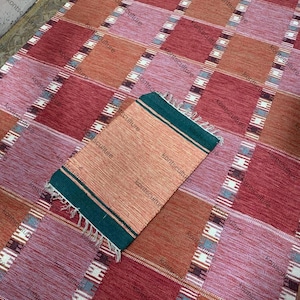 May include: A woven rug with a checkered pattern in shades of red, pink, and orange. A smaller matching rug with a green border and fringe is placed on top. The rugs are made of a textured material.