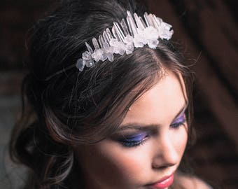May include: A woman with long brown hair wears a crystal and flower crown. The crown is made of clear quartz crystals and white flowers. The woman is wearing blue eyeshadow and has a soft, natural look.