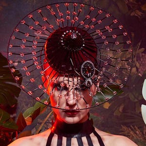May include: A black latex halter top and matching headpiece with a circular design and red accents. The headpiece has a central dome and a decorative spider-like embellishment. The background features lush green foliage.
