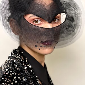 Puede incluir: Una persona con una m&aacute;scara facial de malla negra con un detalle circular de malla blanca. La persona lleva una chaqueta de lentejuelas negra y una camisa negra con cuello. La persona tiene el pelo oscuro y l&aacute;piz labial oscuro.