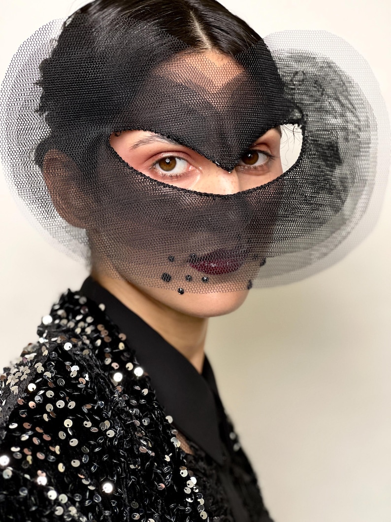 Puede incluir: Una persona con una m&aacute;scara facial de malla negra con un detalle circular de malla blanca. La persona lleva una chaqueta de lentejuelas negra y una camisa negra con cuello. La persona tiene el pelo oscuro y l&aacute;piz labial oscuro.