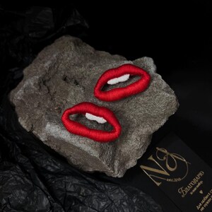 May include: Two embroidered red lips with white teeth are displayed on a gray rock against a black background. The lips are made with red thread, and the teeth are white. The overall theme is fashion and accessories.