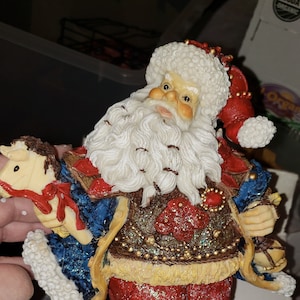 May include: A white, red, and blue Santa Claus figurine holding a rocking horse. He is wearing a blue and gold trimmed coat, red pants, and black boots. The figurine is standing on a snowy base with holly leaves. The base has a barcode sticker that reads "8 05889 01408 3 MADE IN CHINA".