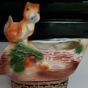 May include: A ceramic planter shaped like a tree stump with a squirrel perched on top. The planter has a brown, green, and white glaze. It has a gold metal stand with three legs.