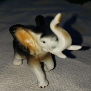 May include: A small porcelain elephant figurine with brown, black, and white paint. The elephant has its trunk raised and is standing on a white surface.