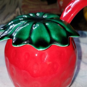 May include: A red ceramic strawberry-shaped sugar bowl with a green leaf lid and a spout. The bowl has a glossy finish and a textured surface.