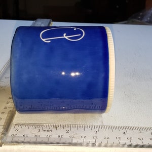 Pu&ograve; includere: Una tazza in ceramica blu con una lettera "G" bianca sul lato. La tazza &egrave; appoggiata su una superficie bianca accanto a un righello.