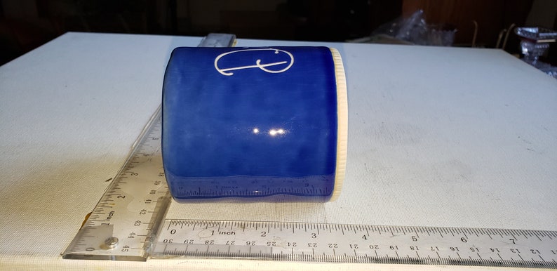 Pu&ograve; includere: Una tazza in ceramica blu con una lettera "G" bianca sul lato. La tazza &egrave; appoggiata su una superficie bianca accanto a un righello.