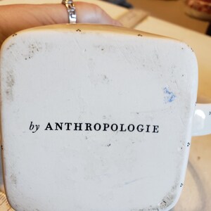 K&ouml;nnte beinhalten: Unteransicht einer wei&szlig;en Keramik-Tasse mit dem Text "by ANTHROPOLOGIE" in Schwarz aufgedruckt. Die Tasse hat einen quadratischen Boden und kleine schwarze Punkte an den R&auml;ndern. Die Tasse wird von einer Person gehalten.