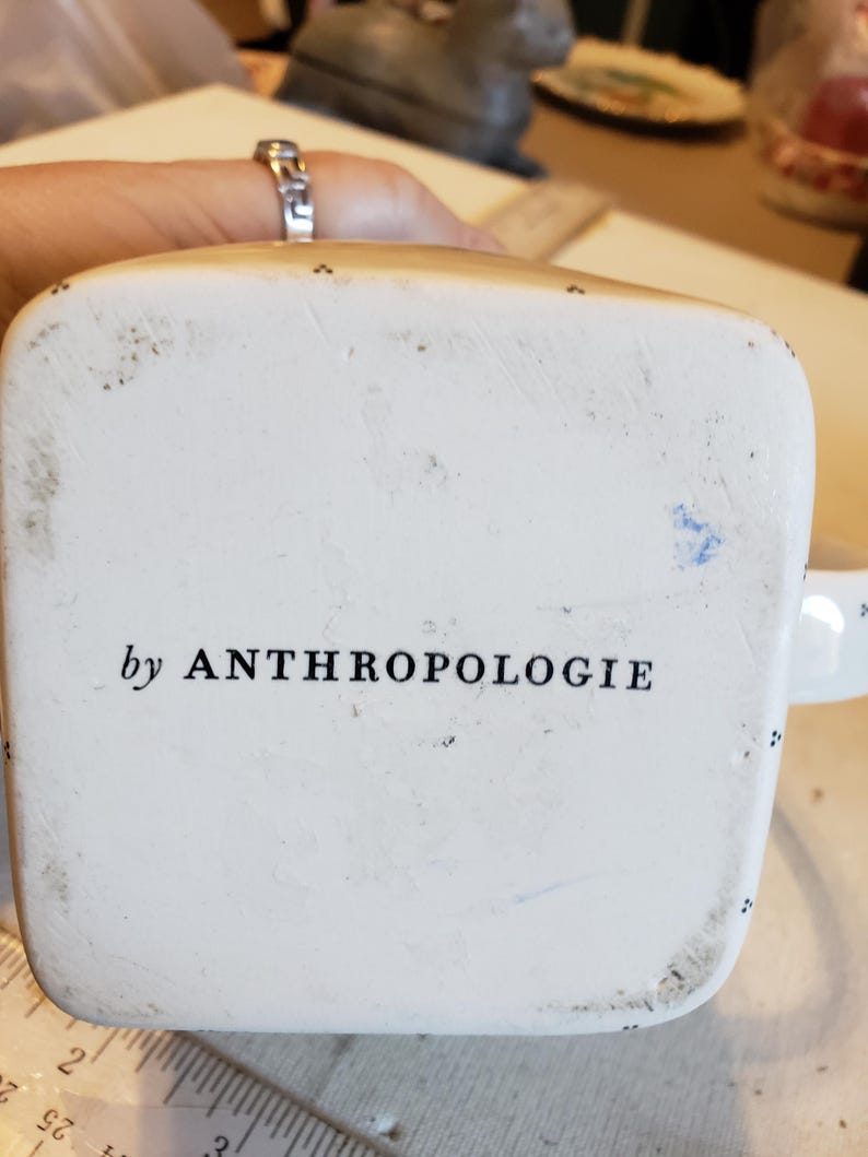 K&ouml;nnte beinhalten: Unteransicht einer wei&szlig;en Keramik-Tasse mit dem Text "by ANTHROPOLOGIE" in Schwarz aufgedruckt. Die Tasse hat einen quadratischen Boden und kleine schwarze Punkte an den R&auml;ndern. Die Tasse wird von einer Person gehalten.