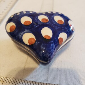 May include: A heart-shaped ceramic trinket box. The box is navy blue with a pattern of white circles, each with a small orange center. The box is approximately 3 inches long.
