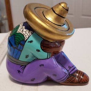 May include: A ceramic figurine of a person wearing a gold sombrero, seated with their head resting on their arms. The figure is painted with vibrant colors, including teal, purple, and brown. The hat is gold, and the figure is wearing purple pants.
