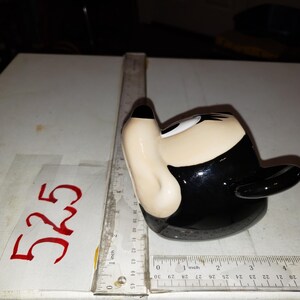 May include: A ceramic figurine shaped like a cartoon character's head, featuring a black hat, black hair, and a beige face. The figurine is on a white surface with a ruler and the number "525" written in red.