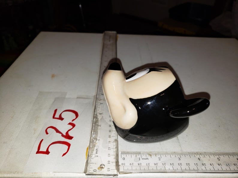 May include: A ceramic figurine shaped like a cartoon character's head, featuring a black hat, black hair, and a beige face. The figurine is on a white surface with a ruler and the number "525" written in red.