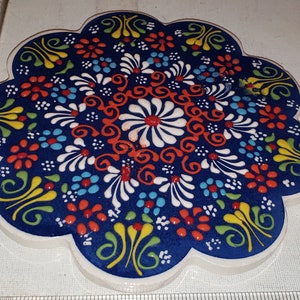 May include: A white ceramic trivet with a blue background and a colorful floral design. The design features red, yellow, green, and white flowers and swirls.