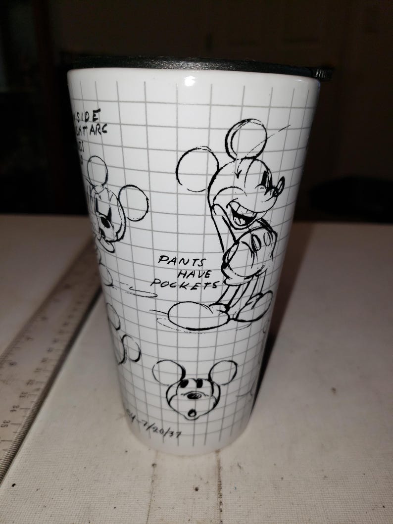 May include: White travel mug featuring a Mickey Mouse sketch design. The mug has a black lid and a grid pattern background. Text includes "Pants Have Pockets" and "Side Ant Arc". The date "7/20/37" is also visible.