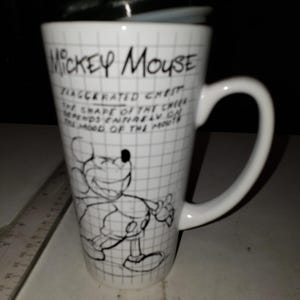 May include: White ceramic travel mug featuring a black and white illustration of Mickey Mouse. The mug has a black lid and a large handle. Text on the mug reads "Mickey Mouse" and a quote about the shape of the cheek.