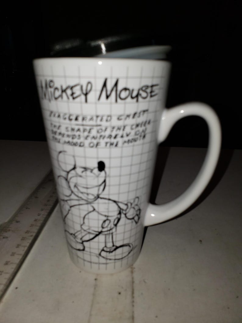 May include: White ceramic travel mug featuring a black and white illustration of Mickey Mouse. The mug has a black lid and a large handle. Text on the mug reads "Mickey Mouse" and a quote about the shape of the cheek.