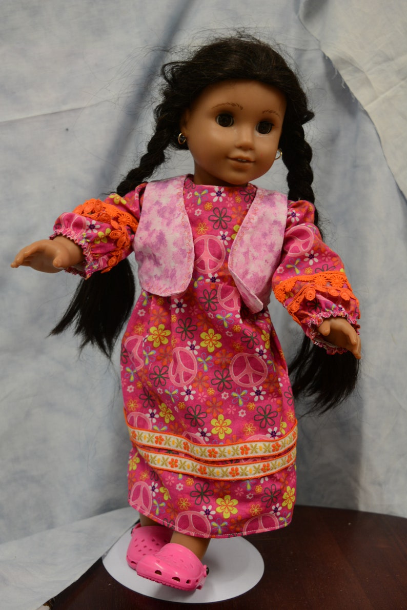 May include: A doll wearing a pink and orange dress with a peace sign pattern and a pink vest. The doll has long black hair and brown eyes. The doll is wearing pink shoes.