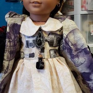 May include: A doll wearing a cream-colored dress with a floral pattern and a collared jacket with a skull and apothecary print. The doll has dark hair, gold hoop earrings, and a black pendant.