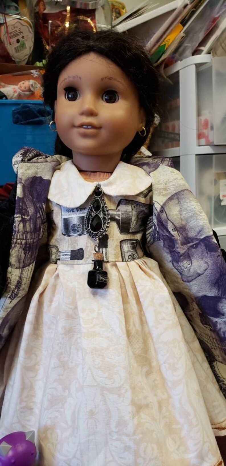 May include: A doll wearing a cream-colored dress with a floral pattern and a collared jacket with a skull and apothecary print. The doll has dark hair, gold hoop earrings, and a black pendant.