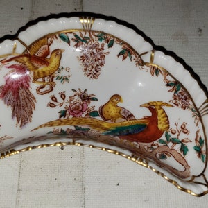 May include: A white porcelain half-moon shaped dish with a gold rim and a colorful floral and bird design. The design features three birds, two yellow and one red, with green and brown foliage.