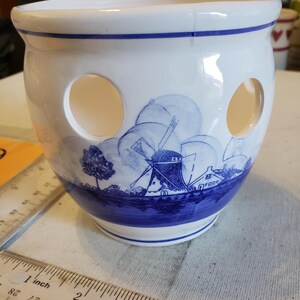 May include: A white ceramic bulb planter with a blue and white windmill design. The planter has three circular openings and a blue band around the top and base. A ruler is in the foreground.