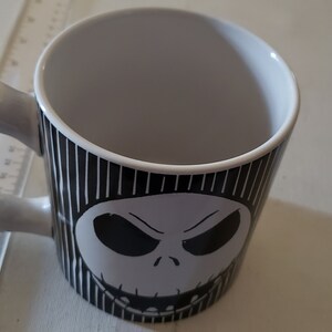 May include: A white ceramic mug with a black and white striped design featuring a black and white illustration of Jack Skellington's face from the Nightmare Before Christmas.