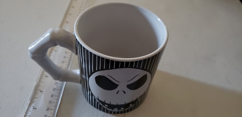 May include: A white ceramic mug with a black and white striped design featuring a black and white illustration of Jack Skellington's face from the Nightmare Before Christmas.