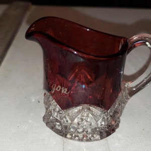 May include: A small, clear glass creamer with a ruby red glass insert. The creamer has a cut glass design and the words "Oregon" are etched on the side.