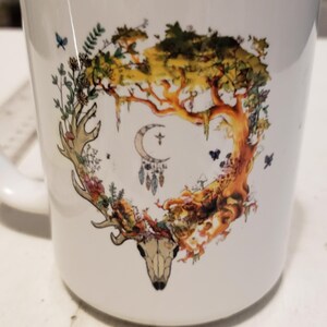 May include: White ceramic mug with a colorful design featuring a deer skull surrounded by a tree, flowers, and butterflies. The design also includes a crescent moon and a dreamcatcher.