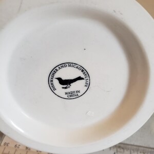 以下が含まれることがあります： 黒い鳥のロゴと「Dishwasher and Microwave Safe」と「Made in China」の文字が入った白いセラミックマグの底。画像の下部には定規が見えます。