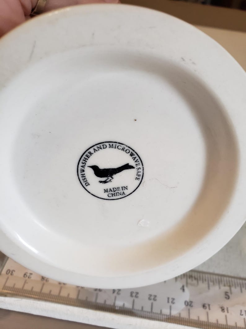 以下が含まれることがあります： 黒い鳥のロゴと「Dishwasher and Microwave Safe」と「Made in China」の文字が入った白いセラミックマグの底。画像の下部には定規が見えます。