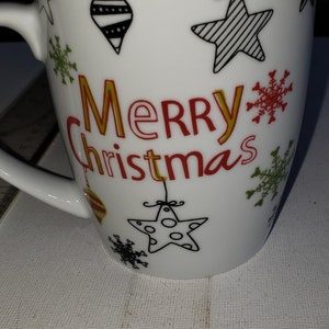May include: White ceramic mug with a black rim and a festive design. The mug features a red and gold "Merry Christmas" message, along with black and white illustrations of stars, snowflakes, and ornaments.
