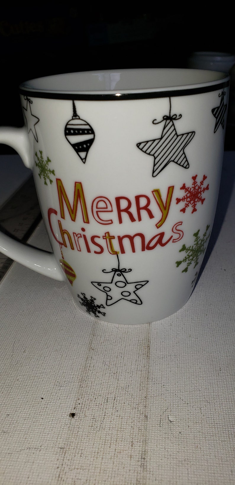 May include: White ceramic mug with a black rim and a festive design. The mug features a red and gold "Merry Christmas" message, along with black and white illustrations of stars, snowflakes, and ornaments.