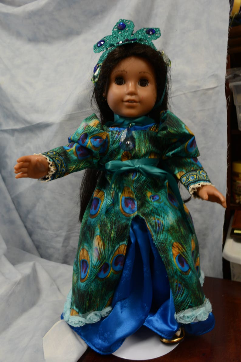 May include: A doll wearing a blue and green peacock-patterned dress with a blue satin skirt and a green beaded crown.