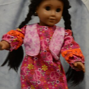 May include: A doll with long black braids wearing a pink vest over a pink and orange dress with peace signs and flowers. The doll has brown eyes and is wearing earrings.