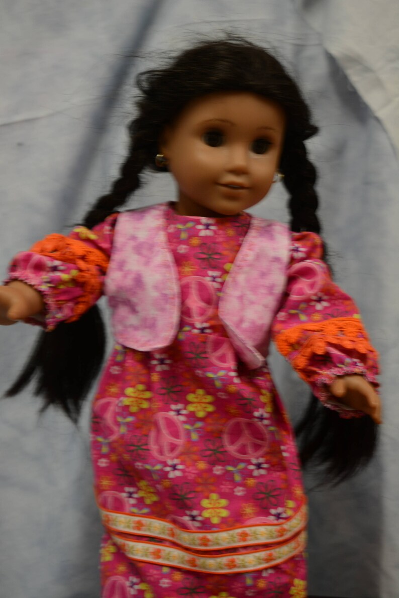 May include: A doll with long black braids wearing a pink vest over a pink and orange dress with peace signs and flowers. The doll has brown eyes and is wearing earrings.